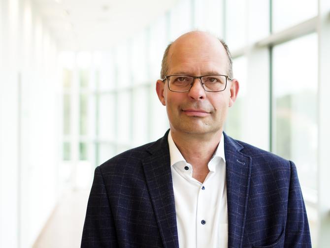 Portrait von Karsten Mueller-Janssen in Anzug und weißem Hemd. Im Hintergrund ist eine Fensterscheibenfront.