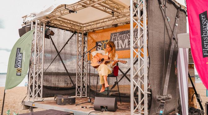 Eine Singer-Songwriterin sitzt barfuß mit einer Akustikgitarre auf einer überdachten Strandbühne beim Flensburger Sommer Open-Air. Links weht eine grüne Flagge und im Hintergrund hängt ein Banner des 'Flensburger Sommer' Festivals direkt am Ostseestrand.