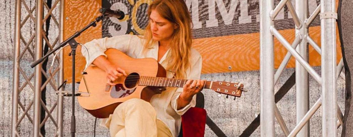 Eine Singer-Songwriterin sitzt barfuß mit einer Akustikgitarre auf einer überdachten Strandbühne beim Flensburger Sommer Open-Air. Links weht eine grüne Flagge und im Hintergrund hängt ein Banner des 'Flensburger Sommer' Festivals direkt am Ostseestrand.