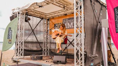 Eine Singer-Songwriterin sitzt barfuß mit einer Akustikgitarre auf einer überdachten Strandbühne beim Flensburger Sommer Open-Air. Links weht eine grüne Flagge und im Hintergrund hängt ein Banner des 'Flensburger Sommer' Festivals direkt am Ostseestrand.