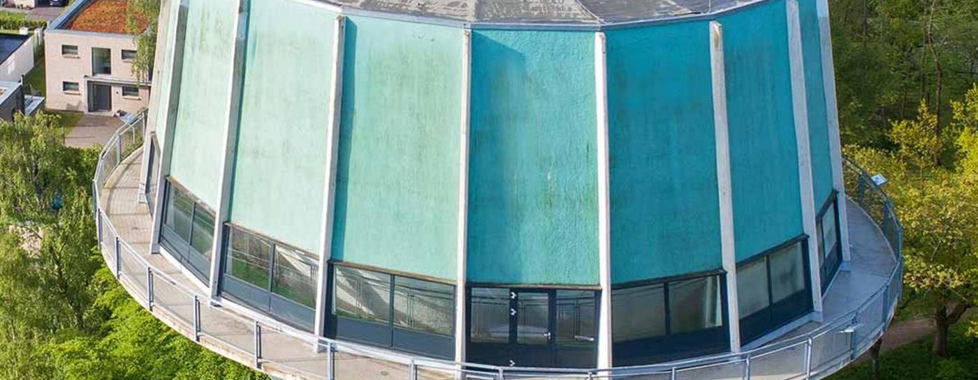 Blick von oben auf den hellgrünen Wasserturm Flensburg, umgeben von Bäumen und modernen Mehrfamilienhäusern. 