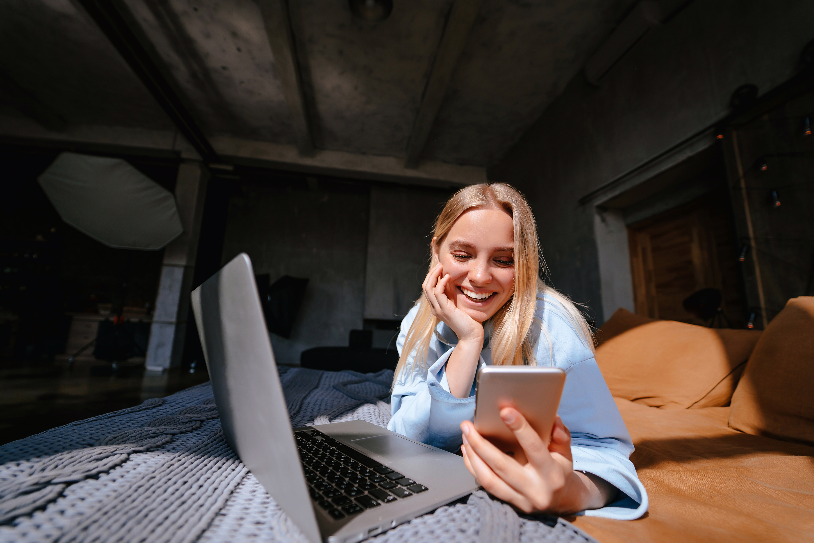 Eine junge Frau mit blonden Haaren und einer blauen Bluse liegt auf einem Bett und schaut lächelnd auf ihr Handy. Vor ihr liegt zusätzlich ein aufgeklappter Laptop. Der Hintergrund ist dunkel gehalten, einige Wände und Gegenstände sind wahrzunehmen.