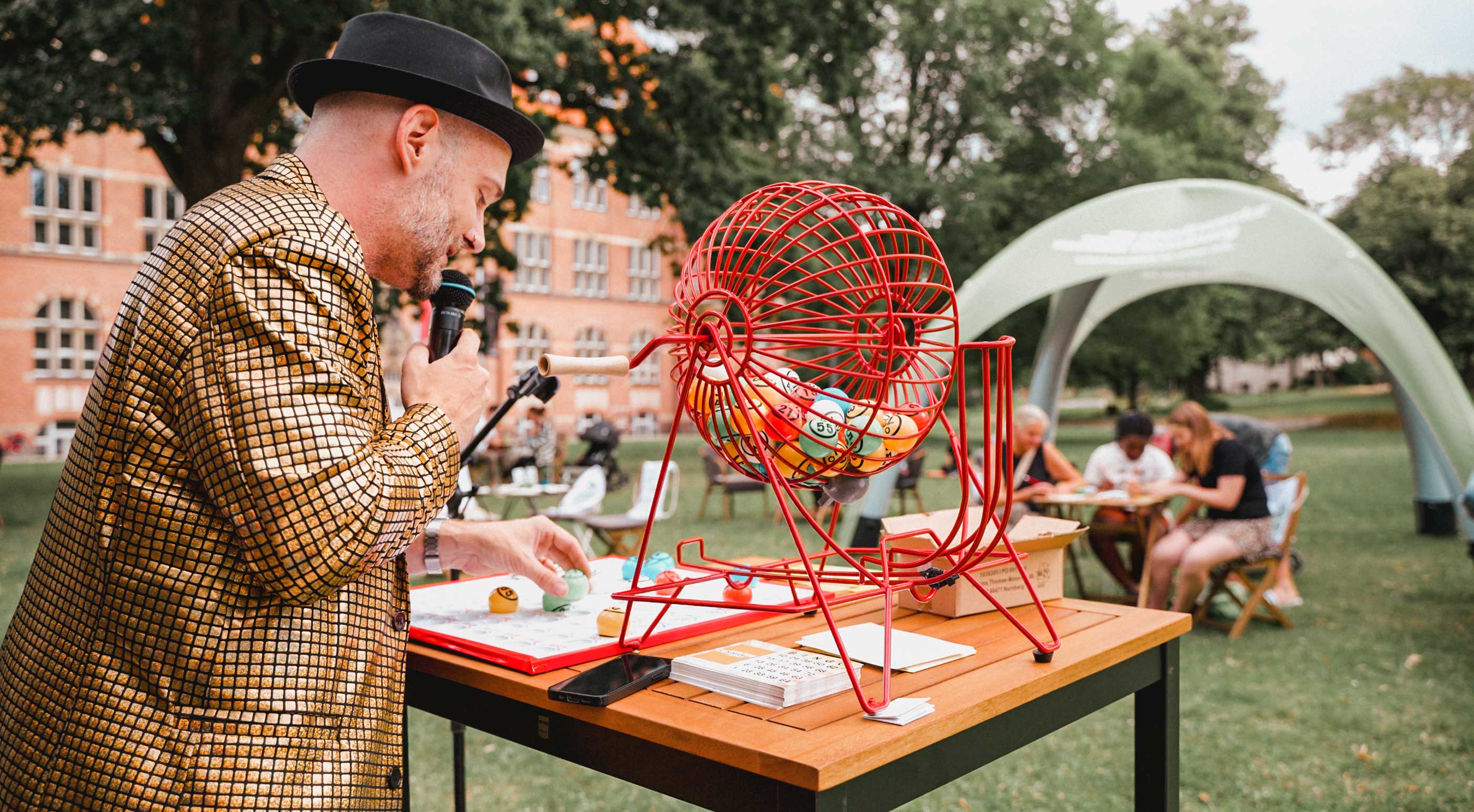 Ein Moderator im goldenen Glitzer-Sakko und schwarzem Hut zieht Bingo-Kugeln aus einer großen, roten Lostrommel und spricht in ein Mikrofon. Im Hintergrund spielen Menschen an Tischen im Park Bingo, nahe einem Backsteingebäude beim Flensburger Sommer Event.