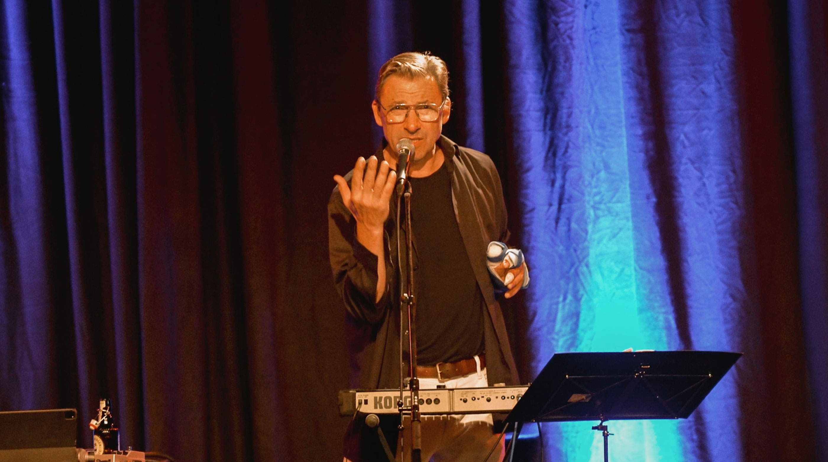Ein Künstler mit Brille spricht gestikulierend in ein Mikrofon auf einer Eventbühne. Er steht vor einem Keyboard (Korg) und einem Notenständer vor einem dunkelblauen, stimmungsvoll beleuchteten Bühnenvorhang.