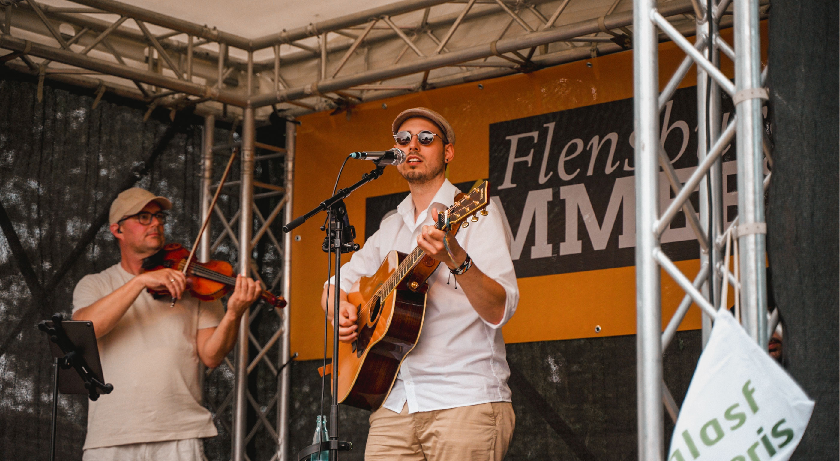 Ein Sänger mit Akustikgitarre, Sonnenbrille und Schiebermütze singt live auf einer Bühne beim Flensburger Sommer Open-Air. Links neben ihm spielt ein Musiker mit Cappy Geige. Im Hintergrund ist ein gelbes Banner mit der Aufschrift 'Flensburger Sommer' zu sehen.