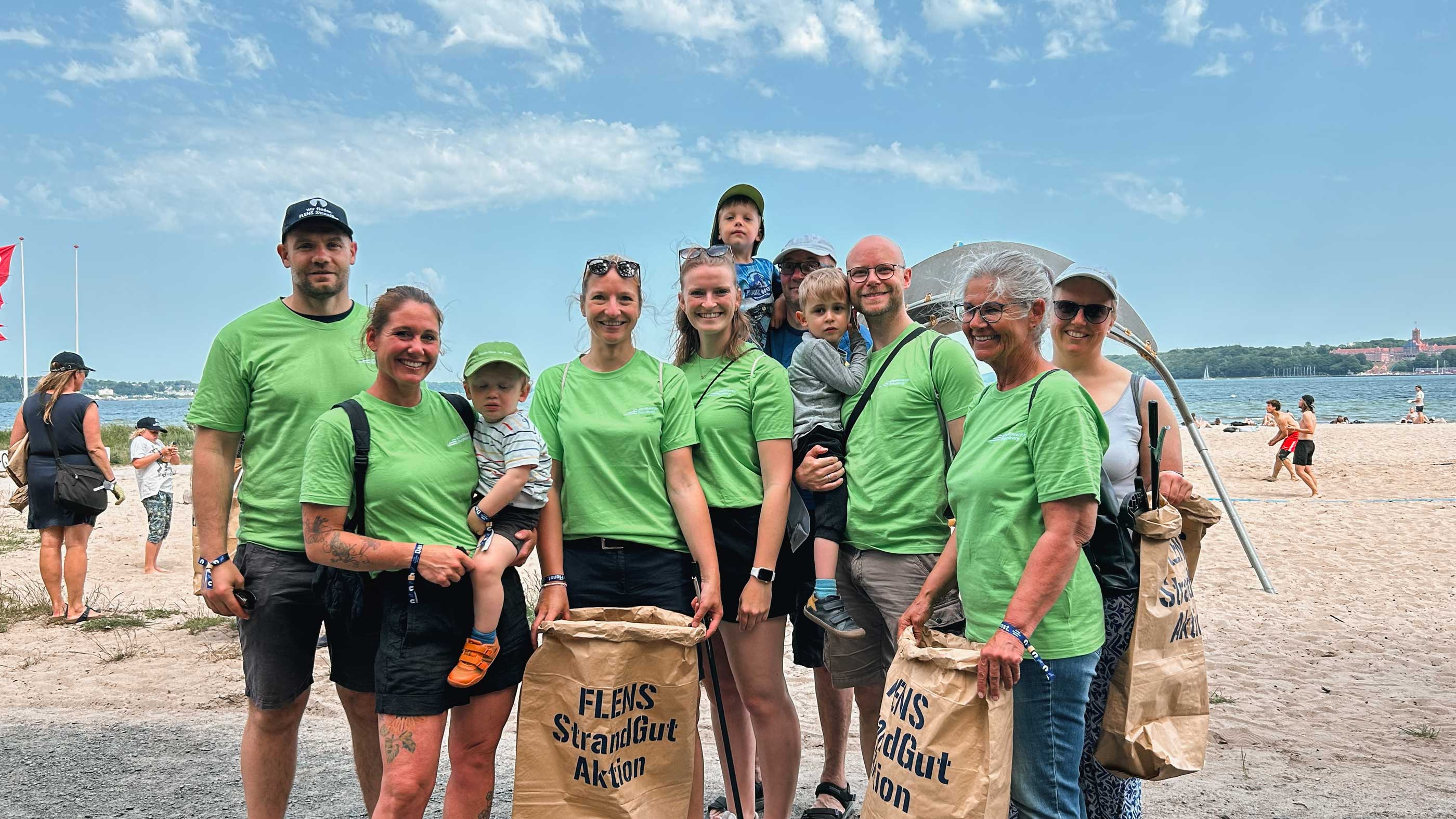 Eine Gruppe von Freiwilligen, Erwachsenen und Kinder, lächelt am Strand nach einer Strandreinigungsaktion. Die Teilnehmer tragen grüne T-Shirts und halten braune Papiertüten mit dem Aufdruck "FLENS StrandGut Aktion". Im Hintergrund ist die Flensburger Förde zu sehen.